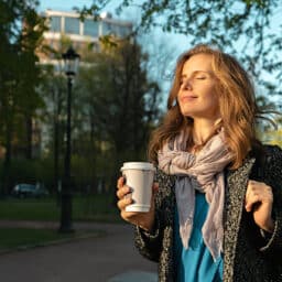 Woman holding a coffee cup standing outside on a beautiful spring day, enjoying the warmth and the sunshine on her face.