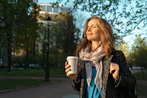 Woman holding a coffee cup standing outside on a beautiful spring day, enjoying the warmth and the sunshine on her face.