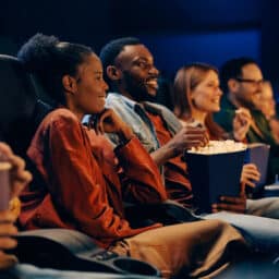 Focus is on a young couple sharing popcorn with additional movie goers seated to either side of them.