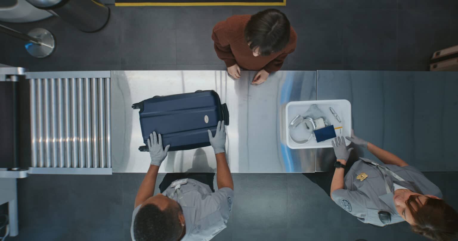 Overhead view of going through airport security; agent handles a carry-on suitcase on the conveyor belt.